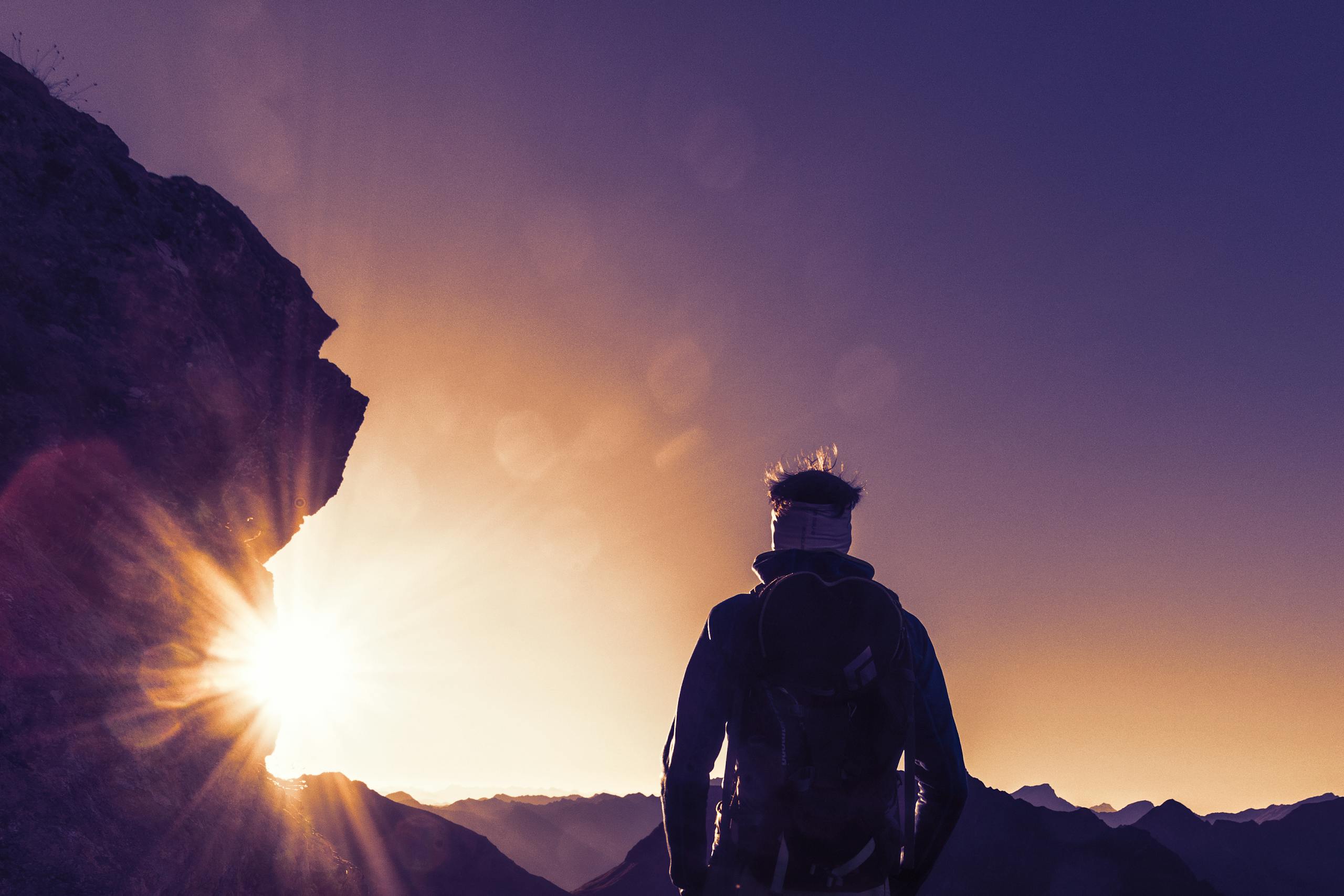 Silhouette of a hiker climbing up a mountain trail at sunrise with sun flares and mountains in the background.
