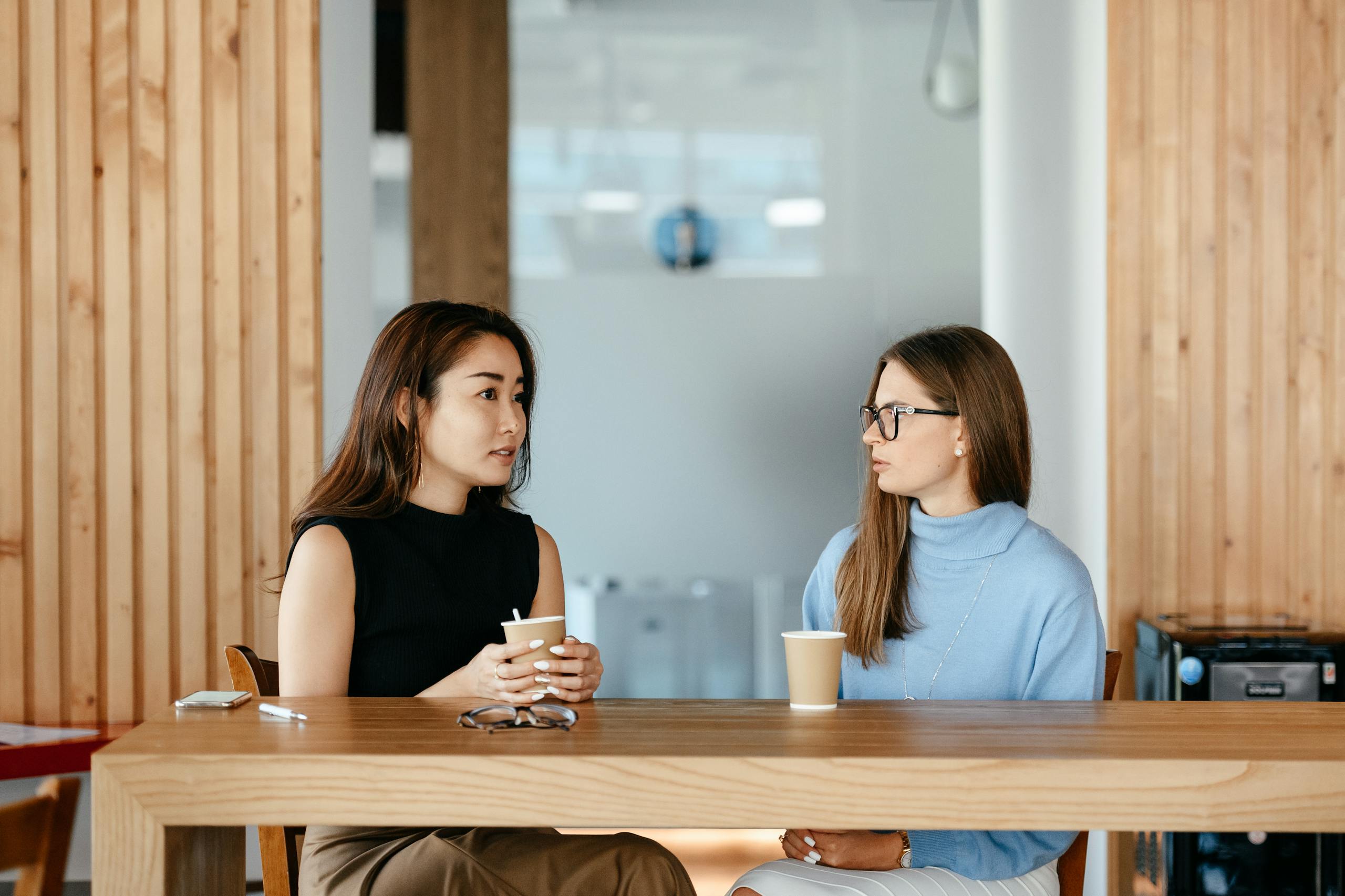 Two women enjoy a coffee and chat at a modern indoor cafe setting.